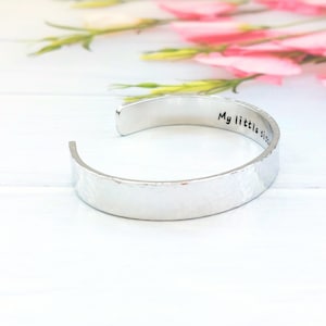 May include: A silver cuff bracelet with a textured finish. The open bracelet has the words "My little sister" engraved inside. Pink flowers are visible in the background.