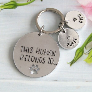 May include: Silver keyring with a round tag that says "This human belongs to..." with a paw print cutout. Two smaller round tags with paw prints and the names "Rosie" and "Cleo" are attached to the keyring.