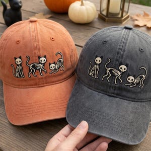 May include: Two baseball caps, one orange and one charcoal gray, each featuring an embroidered design of three black cat skeletons in different poses. The caps have a structured crown, a curved brim, and a button on top of the fabric dome.