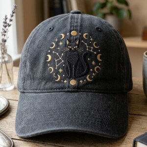 May include: A charcoal gray baseball cap featuring an embroidered design of a black cat centered within a circular arrangement of gold-colored moon phases, stars, and constellations. The cap has a structured crown and a curved brim.