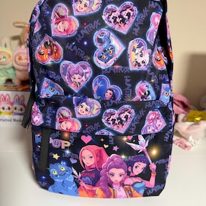 Może przedstawiać: Czarny plecak z kolorowym wzorem w stylu anime. Posiada grafiki w kształcie serca z ilustracjami postaci i powtarzającym się słowem "HUNTR". Dolna kieszeń przedstawia grupę postaci z anime i słowo "POP".