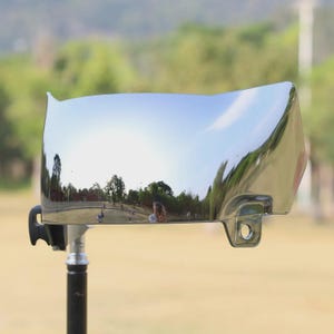 May include: A chrome-finished motorcycle windshield, reflecting a park scene with trees and sky. The windshield has a curved shape with a small lip at the top and a mounting bracket.