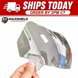 May include: A clear, curved, semi-transparent plastic visor shield with a contoured shape and a mounting tab at the base. The surface is reflective, showing light reflections and a distorted view of the surroundings. The Maxshield logo is visible.