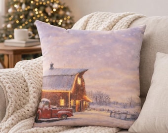 Può includere: Un cuscino decorativo con una scena invernale con un fienile e un camion rossi nella neve. Il cuscino ha un cielo morbido, viola chiaro e rosa con neve che cade. Il fienile ha finestre illuminate e il camion è parcheggiato davanti.