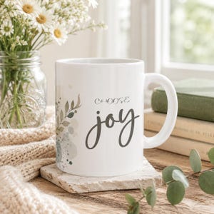 May include: A white ceramic mug featuring the text "CHOOSE joy" in a dark gray script and print font. The mug is decorated with a watercolor-style illustration of green leaves and soft blue-gray circular accents on the left side.