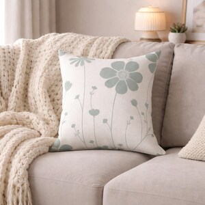 Può includere: Un cuscino decorativo quadrato con un motivo floreale. Il cuscino presenta uno sfondo bianco con un motivo di fiori e steli azzurri. È posto su un divano beige, con una coperta lavorata a maglia color crema visibile sullo sfondo.