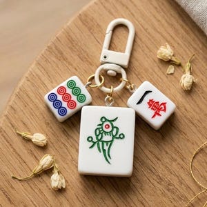 Puede incluir: Un llavero con tres fichas de mahjong blancas unidas a un mosquet&oacute;n de pl&aacute;stico blanco. La ficha central muestra un dise&ntilde;o de p&aacute;jaro verde, mientras que las dos fichas laterales m&aacute;s peque&ntilde;as presentan patrones circulares y caracteres chinos.