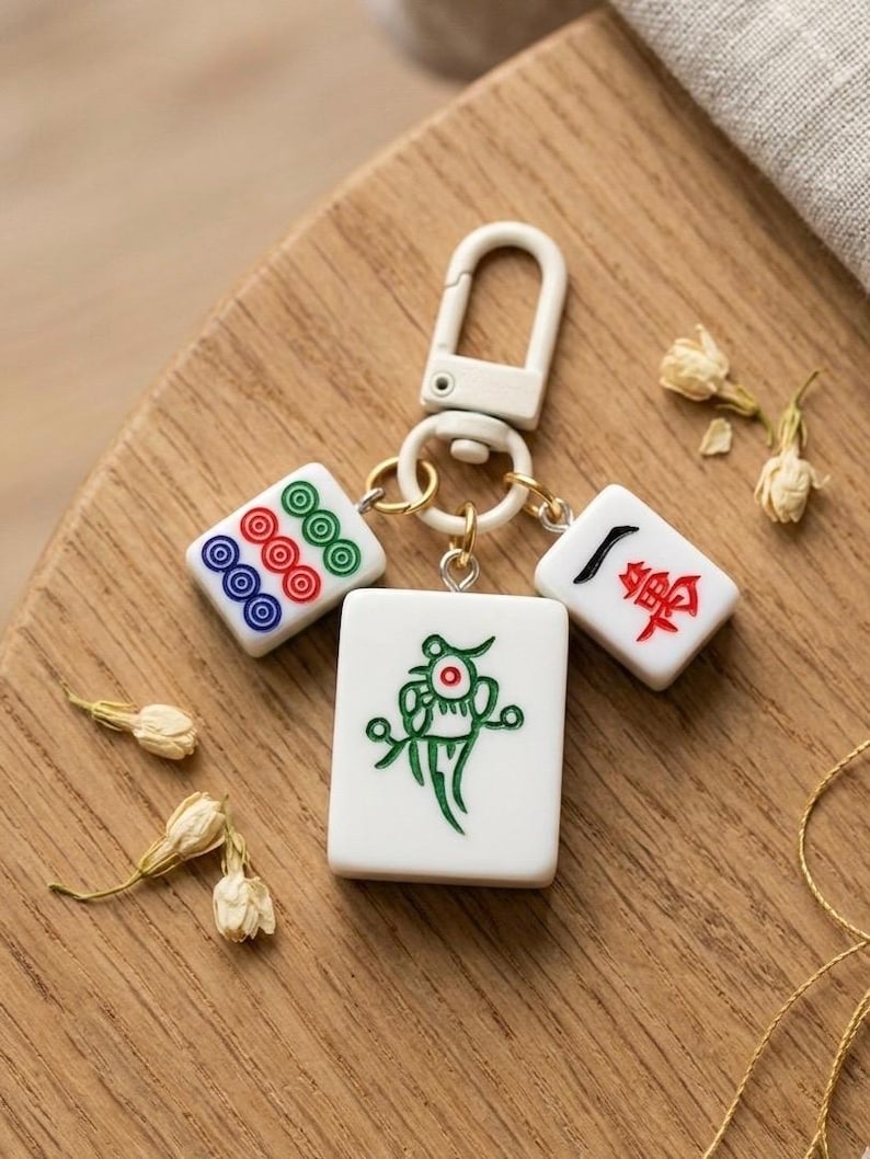 Puede incluir: Un llavero con tres fichas de mahjong blancas unidas a un mosquet&oacute;n de pl&aacute;stico blanco. La ficha central muestra un dise&ntilde;o de p&aacute;jaro verde, mientras que las dos fichas laterales m&aacute;s peque&ntilde;as presentan patrones circulares y caracteres chinos.