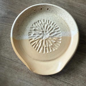 Może przedstawiać: Beżowa ceramiczna podstawka pod łyżkę z teksturowanym, kwiatowym wzorem pośrodku. Obręcz jest nakrapiana, a z boku znajduje się mały dzióbek. Dwa małe otwory znajdują się blisko obręczy. Średnica ok. 20 cm.