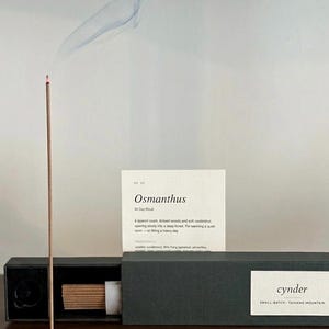 May include: A burning incense stick in a gold-toned metal holder next to a black box containing incense sticks. A white card with the text Osmanthus, All Day Ritual, and a list of ingredients is positioned behind the box. The box is labeled cynder.
