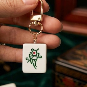 Puede incluir: Un llavero rectangular de ficha de mahjong blanca con un dise&ntilde;o grabado en verde y un peque&ntilde;o acento rojo en el centro. La ficha est&aacute; unida a un mosquet&oacute;n y una anilla giratoria de metal dorado para sujetarla a llaves o bolsos.