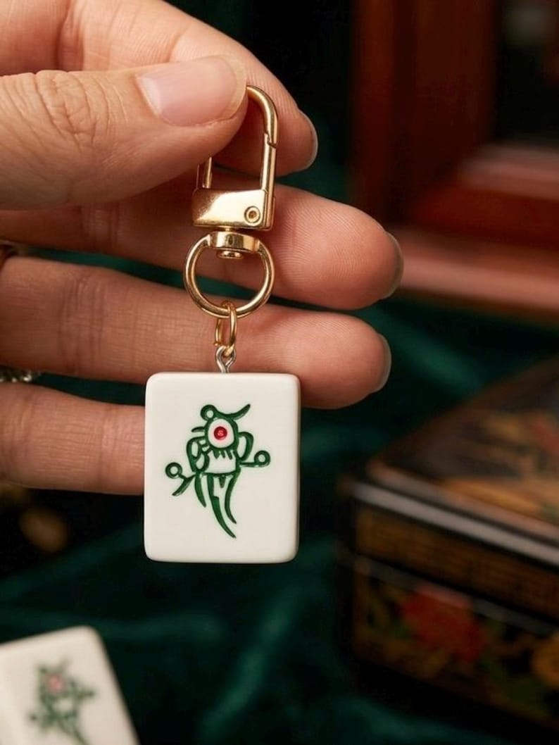 Puede incluir: Un llavero rectangular de ficha de mahjong blanca con un dise&ntilde;o grabado en verde y un peque&ntilde;o acento rojo en el centro. La ficha est&aacute; unida a un mosquet&oacute;n y una anilla giratoria de metal dorado para sujetarla a llaves o bolsos.