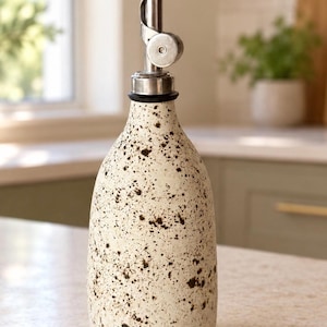 Può includere: Una bottiglia dosatrice per olio in ceramica color crema con un motivo a macchie marrone scuro su tutto l'esterno. La bottiglia è sormontata da un beccuccio in metallo argentato che include un coperchio incernierato e una guarnizione nera.