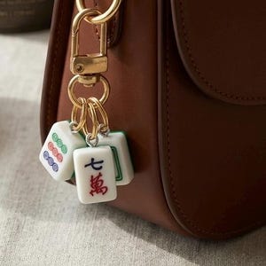 Puede incluir: Un bolso de cuero marr&oacute;n con un llavero de mosquet&oacute;n de metal dorado. El llavero sostiene tres fichas de mahjong blancas, incluida una con el car&aacute;cter chino rojo para siete y dos fichas que muestran patrones circulares en verde, rojo y azul.
