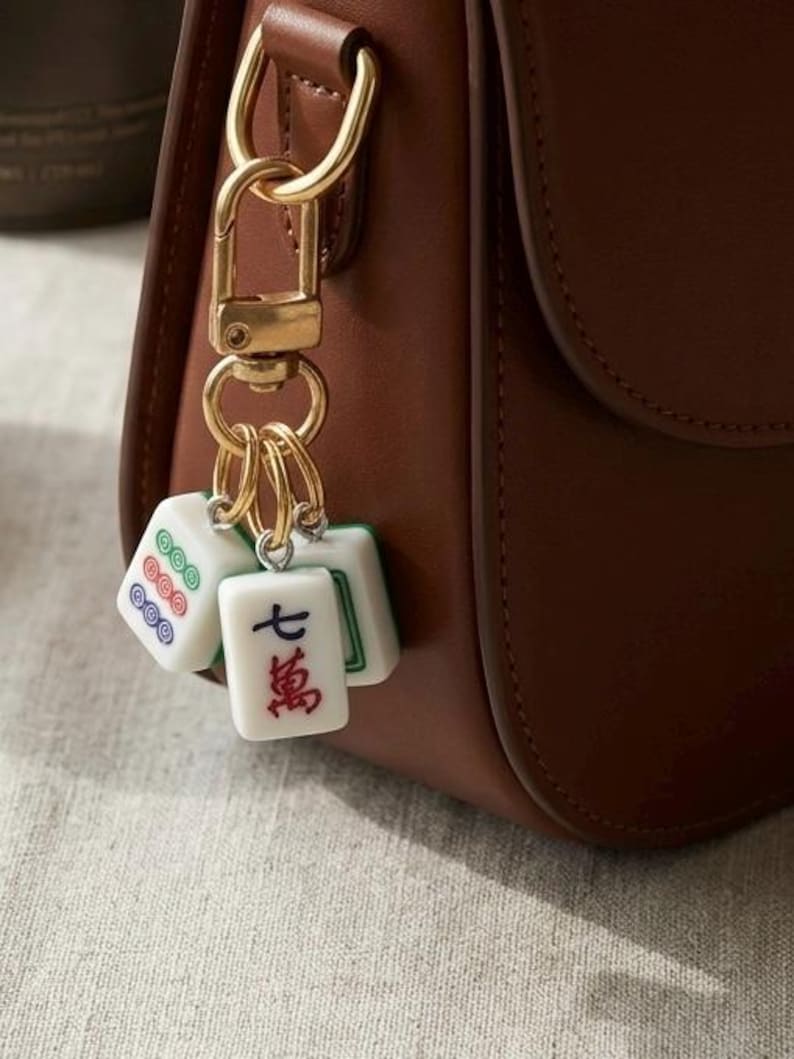 Puede incluir: Un bolso de cuero marr&oacute;n con un llavero de mosquet&oacute;n de metal dorado. El llavero sostiene tres fichas de mahjong blancas, incluida una con el car&aacute;cter chino rojo para siete y dos fichas que muestran patrones circulares en verde, rojo y azul.