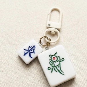 Puede incluir: Un llavero con dos fichas de mahjong rectangulares blancas unidas a un cierre giratorio de pl&aacute;stico color crema. Una ficha muestra un car&aacute;cter chino azul, mientras que la m&aacute;s grande presenta un gr&aacute;fico verde con un punto rojo central.