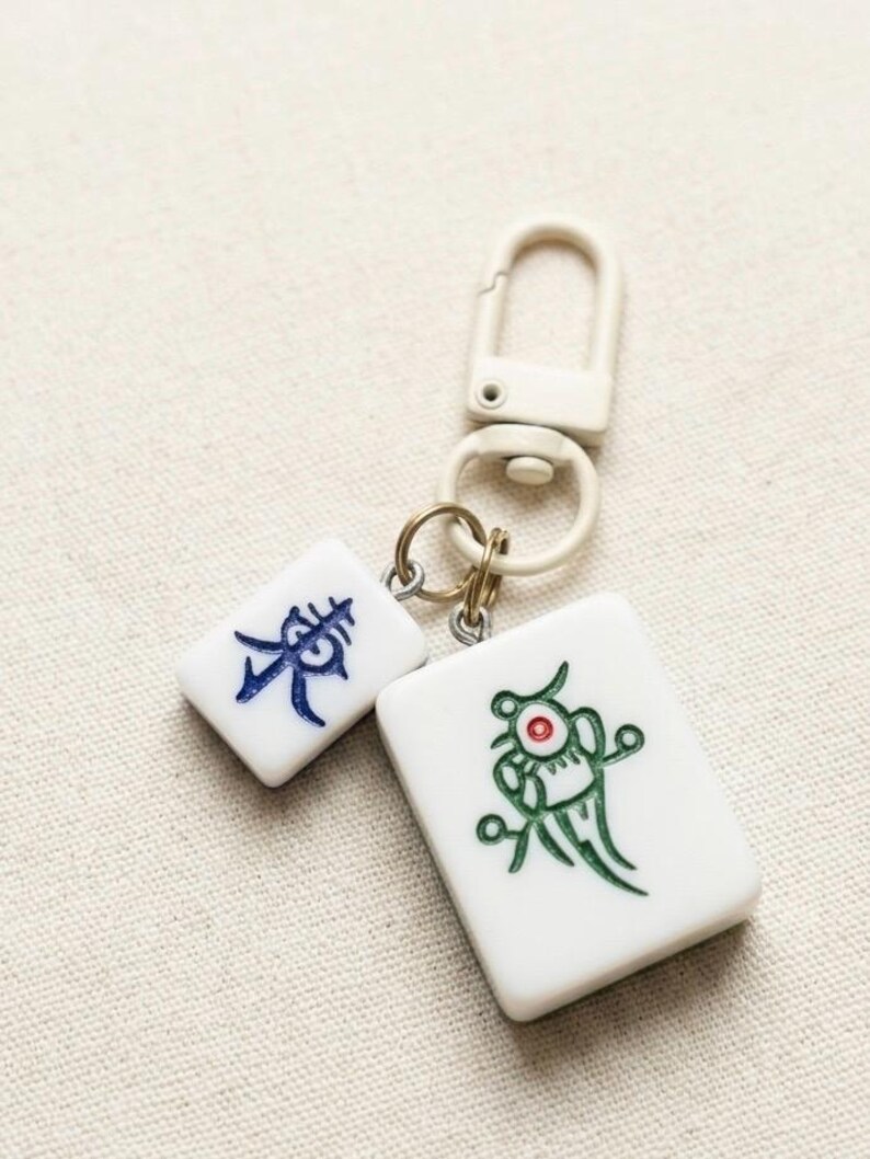 Puede incluir: Un llavero con dos fichas de mahjong rectangulares blancas unidas a un cierre giratorio de pl&aacute;stico color crema. Una ficha muestra un car&aacute;cter chino azul, mientras que la m&aacute;s grande presenta un gr&aacute;fico verde con un punto rojo central.