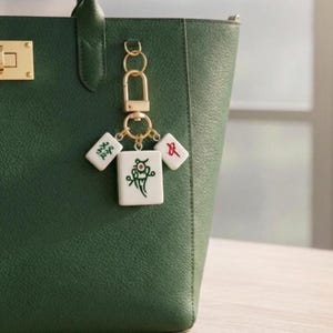 Pode incluir: Um porta-chaves de metal dourado com três peças brancas de mahjong presas a um fecho mosquetão. A peça central apresenta um desenho de pássaro em verde e vermelho, enquanto as duas peças laterais menores exibem caracteres chineses.