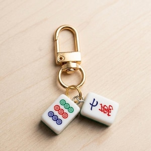 Puede incluir: Un llavero de metal dorado con dos fichas de mahjong rectangulares blancas como dijes. Una ficha muestra tres s&iacute;mbolos circulares en azul, rojo y verde, mientras que la otra tiene un car&aacute;cter azul y uno rojo grabados en su superficie.