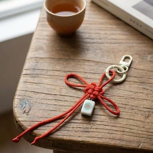 Puede incluir: Un llavero con un cord&oacute;n de color naranja brillante atado en un nudo decorativo, unido a una anilla de metal blanco. Una peque&ntilde;a pieza rectangular blanca con un dise&ntilde;o gr&aacute;fico floral en verde y rojo cuelga del centro del cord&oacute;n anudado.