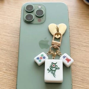 Puede incluir: Un smartphone color verde salvia con un dije dorado en forma de coraz&oacute;n unido a un broche de langosta met&aacute;lico. Tres fichas de mahjong blancas cuelgan del broche, con un dise&ntilde;o de p&aacute;jaro verde, un patr&oacute;n circular rojo y azul y caracteres chinos rojos.