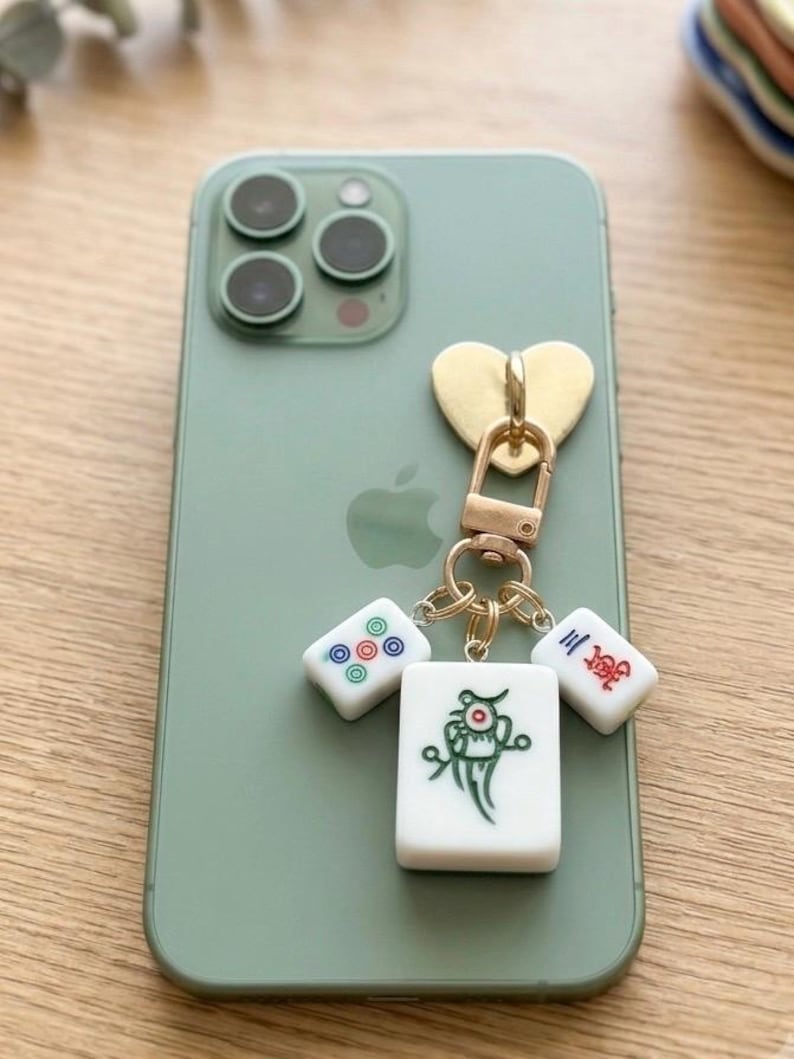 Puede incluir: Un smartphone color verde salvia con un dije dorado en forma de coraz&oacute;n unido a un broche de langosta met&aacute;lico. Tres fichas de mahjong blancas cuelgan del broche, con un dise&ntilde;o de p&aacute;jaro verde, un patr&oacute;n circular rojo y azul y caracteres chinos rojos.