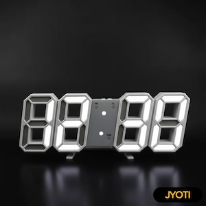 May include: A modern digital clock with large, illuminated white numbers against a gray frame. The clock displays the time in a 3D style, with a dark background. The brand name "JYOTI" is displayed in a black rectangle.