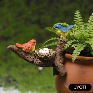 May include: A decorative figurine of two birds, one orange and yellow, the other blue and yellow, on a faux branch with a nest holding three white eggs. The branch is attached to a terracotta pot. The pot has the word "JYOTI".