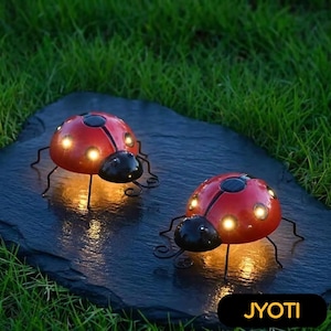 May include: Two red ladybug-shaped solar lights with black accents and glowing lights. The lights are on a dark, textured surface, with green grass in the background. The lights have black legs and antennae.