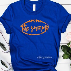 May include: A royal blue t-shirt with an orange football graphic that says "The Swamp" on it. The shirt is made of a soft, comfortable material and is perfect for showing your team spirit.