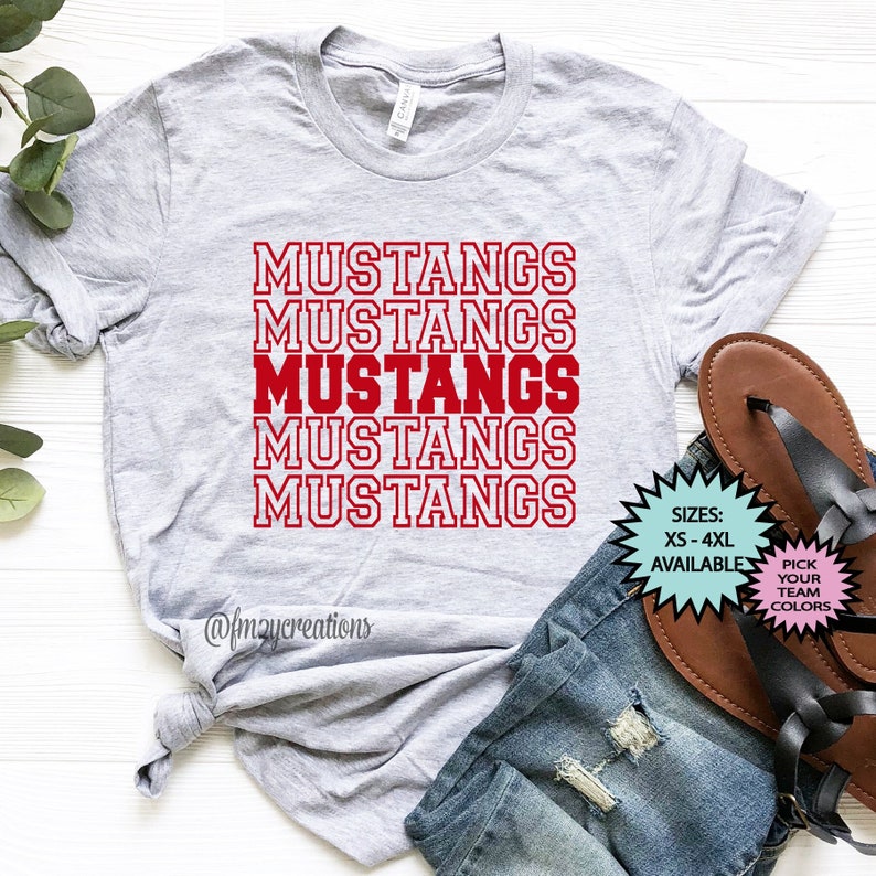 May include: A grey t-shirt with the word "MUSTANGS" repeated five times in red lettering. The shirt is folded and laid on a white surface with a pair of brown sandals and blue jeans. The text "SIZES: XS-4XL AVAILABLE" and "PICK YOUR TEAM COLORS" is also visible in the image.
