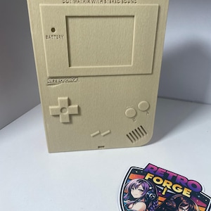 May include: A beige, 3D-printed replica of a retro handheld gaming device, featuring a screen, buttons, and the text "DOT MATRIX WITH STEREO SOUND." The Retroforge logo sticker with anime-style characters is next to the device.