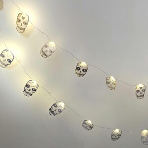 May include: String of decorative skull-shaped lights. Each skull is white with black detailing and a warm, yellow glow. The string lights are strung on a thin, silver wire, perfect for Halloween or gothic-themed decor.