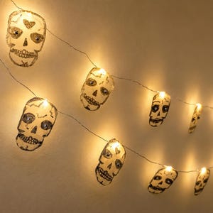 May include: A string of LED fairy lights featuring translucent, flat skull-shaped ornaments. Each skull is decorated with black line art details and emits a warm, glowing yellow light from within, creating a festive illumination effect.