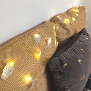 May include: Three decorative pillows. Two pillows are a mustard yellow with a small dot pattern, and the third is a dark gray velvet with embroidered spiders. A string of small skull and ghost lights is draped across the pillows.