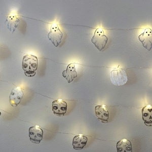 May include: A string of Halloween-themed fairy lights. The lights feature small, illuminated paper cutouts of skulls, ghosts, and a pumpkin. The lights are strung on a thin wire, creating a festive and spooky ambiance.