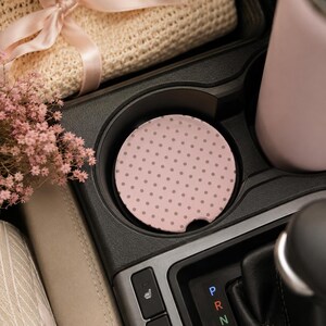 May include: A pink car coaster with a gray polka dot pattern sits in a car's cup holder. The coaster is round with a small notch. A pink tumbler is in the adjacent cup holder. A beige woven item and pink flowers are also visible.