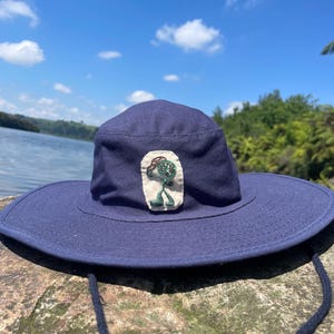 May include: A navy blue sun hat with a wide brim and a chin strap. The hat has a small embroidered patch with a green and red design. The hat is on a rock, with a lake and blue sky in the background.