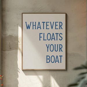 以下が含まれることがあります： 白い背景に青い大文字で「WHATEVER FLOATS YOUR BOAT」と書かれた長方形の額入りサイン。フレームは明るい茶色です。サインは、テクスチャ加工された灰色の壁に掛けられています。