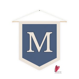 Puede incluir: Un banderín azul marino con un borde color crema y una gran letra "M" color crema en el centro. El banderín está suspendido de un palillo de madera con una cuerda blanca. Un artículo decorativo.