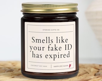 May include: A dark glass jar candle with a gold lid. The label reads "Smells like your fake ID has expired" with "Happy 21st, Olivia!" below. The candle is made with coconut soy wax.