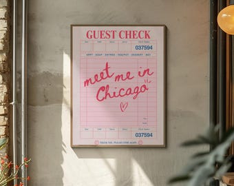 May include: A framed pink guest check print with the words "meet me in Chicago" in a handwritten style. The print includes a check number and a heart symbol. The background is a neutral-colored wall.