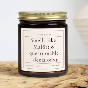 May include: A dark brown glass jar candle with a gold lid. The label reads "Smells like Malört & questionable decisions." The candle is made with coconut soy wax and weighs 255 g.
