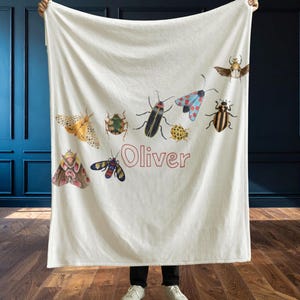 May include: A cream-colored blanket featuring colorful illustrations of various insects, including beetles and moths. The name "Oliver" is printed in a red font. The blanket is held up against a dark blue wall and a wooden floor.