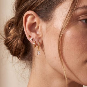 May include: A selection of gold earrings are displayed on an ear. The set includes a small ball stud, a cluster of three small white stones, a chain-linked earring, and a gold hoop with a rectangular, clear stone. The earrings are displayed on a person's ear.