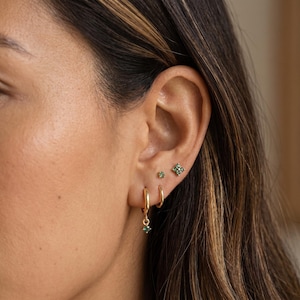May include: A set of gold earrings consisting of a small hoop with a dangling green gemstone, a tiny single green gemstone stud, and a four-petaled flower-shaped stud featuring four small green gemstones set in a gold metal frame.