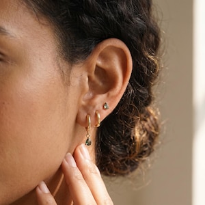 May include: A set of gold jewelry consisting of two small hoop earrings and one stud earring. The stud and one hoop feature a small, teardrop-shaped green gemstone. The jewelry is worn in the earlobe, with the hoops stacked and the stud above.