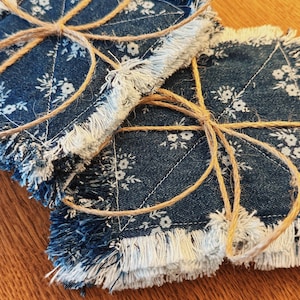 May include: Two square denim coasters with white floral patterns and frayed edges, tied together with twine. The coasters are a dark blue color and are likely handmade. The background is a wooden surface.