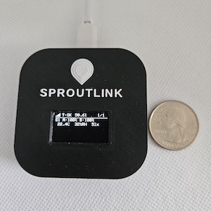 Peut inclure: Un appareil Sproutlink noir avec un petit écran affichant des relevés. L'appareil est carré avec des coins arrondis et un logo blanc. Une pièce de 25 centimes est à côté de l'appareil pour la comparaison de taille. Un câble USB blanc est connecté.