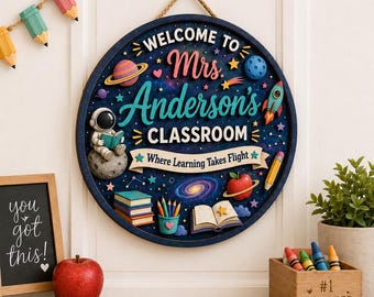 Puede incluir: Un letrero de pared circular de madera con temática espacial y el texto "WELCOME TO Mrs. Anderson's CLASSROOM Where Learning Takes Flight". El diseño incluye un astronauta, planetas, un cohete, libros, lápices y una manzana sobre un fondo azul oscuro.